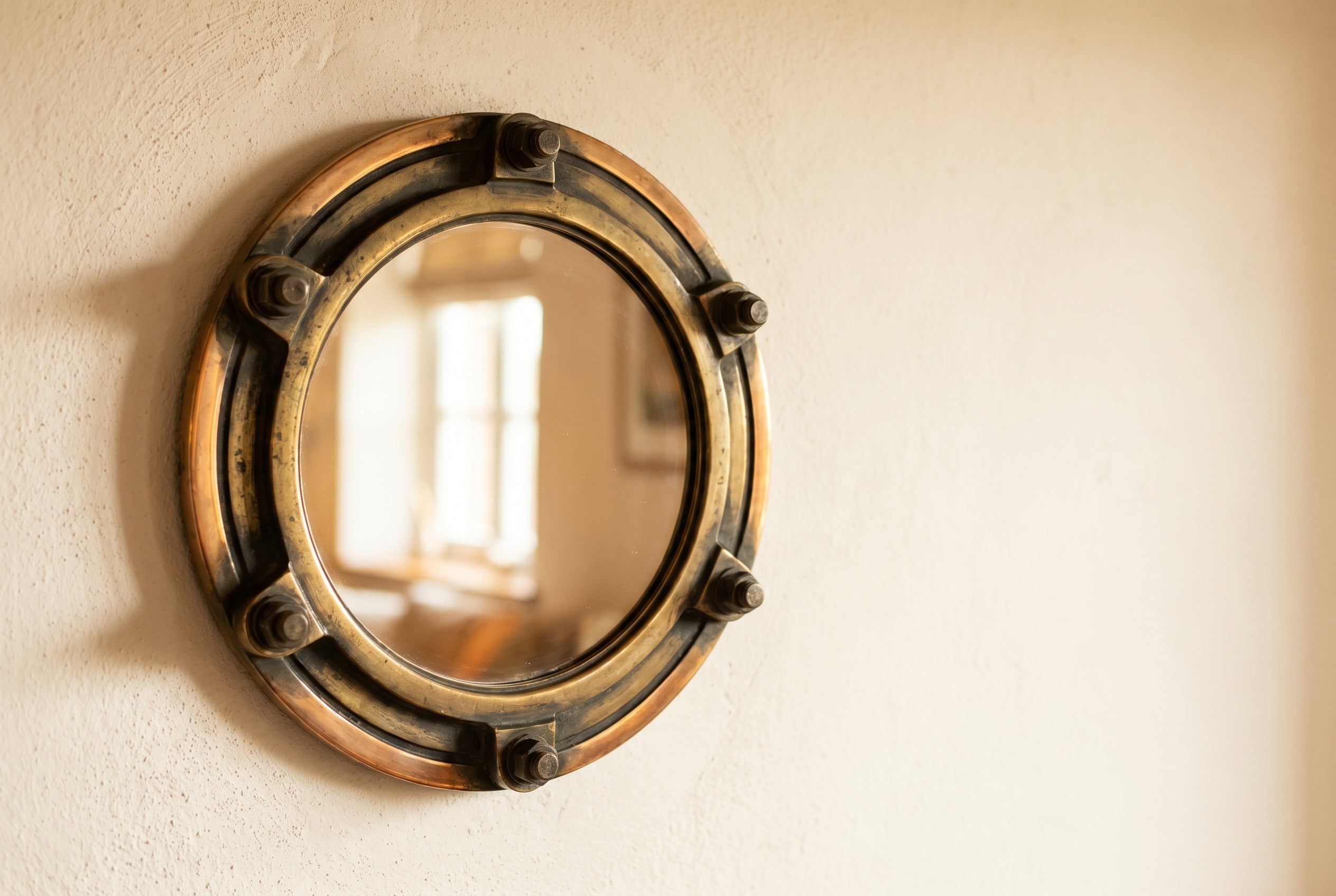 Aged brass-framed circular ship porthole mirror mounted on a warm cream plaster wall, with patina and visible mounting bolts.