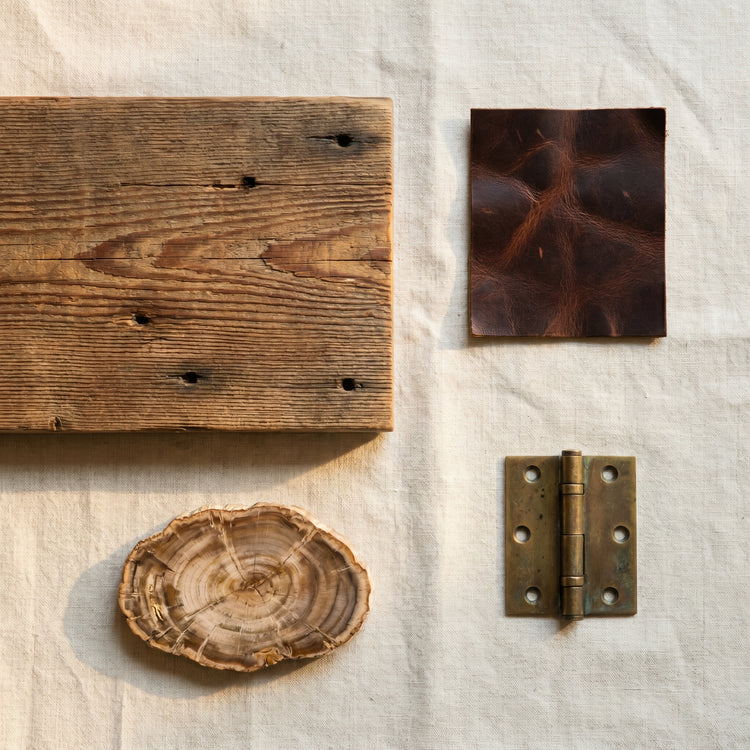 Flat-lay of heritage materials on linen: reclaimed pine, antique buffalo leather, aged brass hinge, petrified wood slice